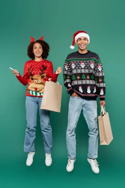 Happy african american couple in christmas outfit levitating with cellphone and shopping bags on green background — Stock Photo