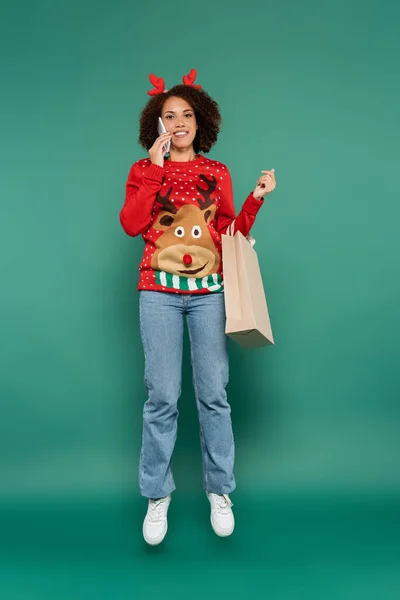 African american woman in christmas outfit talking on smartphone while levitating with shopping bag on green background — Stock Photo