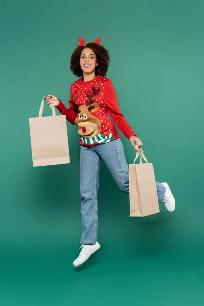 Smiling african american woman in christmas outfit levitating with shopping bags on green background — Stock Photo