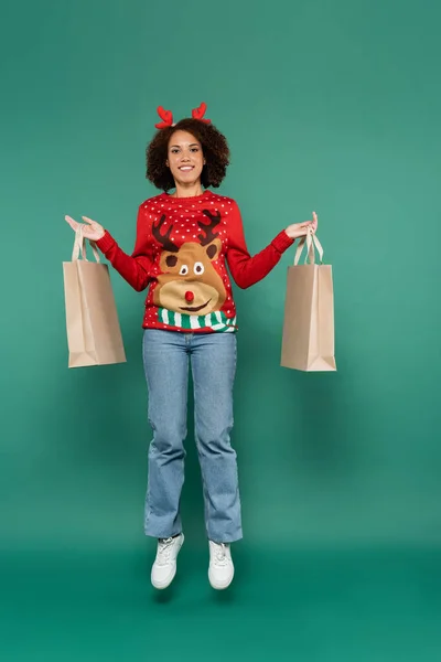 Glücklich afrikanisch-amerikanische Frau im Pullover mit Hirsch und roten Hörnern Stirnband schwebt mit Einkaufstaschen auf grünem Hintergrund — Stockfoto