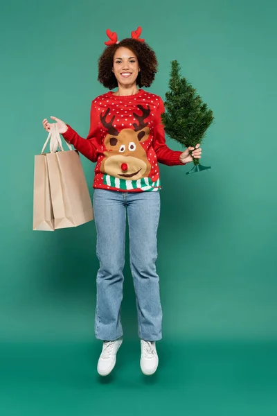 Mujer afroamericana en traje festivo levitando con bolsas de compras y pequeño árbol de Navidad sobre fondo verde - foto de stock