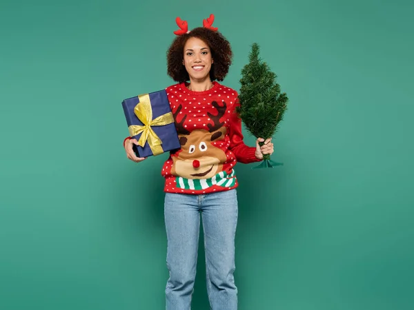 Feliz mulher afro-americana em camisola vermelha com renas segurando árvore de natal e caixa de presente azul isolado no verde — Fotografia de Stock