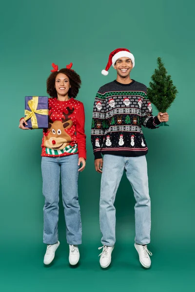 Glückliches afrikanisch-amerikanisches Paar in festlichen Pullovern schwebt mit Geschenkbox und kleinem Weihnachtsbaum auf grünem Hintergrund — Stockfoto
