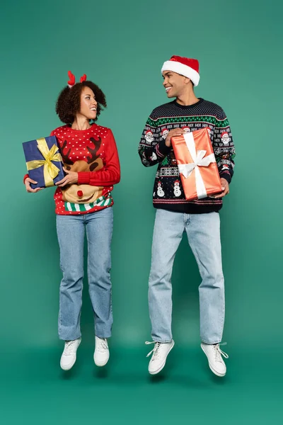 Cheerful african american couple in sweaters with christmas pattern levitating with gift boxes on green background — Stock Photo