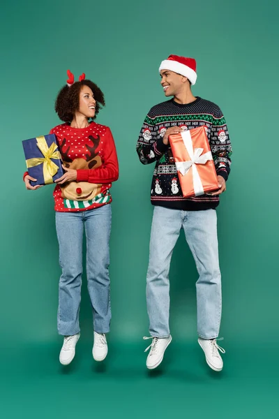 Alegre casal afro-americano em camisolas com padrão de natal levitando com caixas de presente em fundo verde — Fotografia de Stock