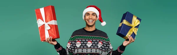 Allegro uomo afroamericano in cappello di Babbo Natale con scatole regalo rosse e blu isolate su verde, banner — Foto stock