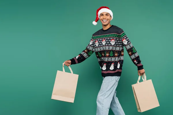 Hombre afroamericano feliz en sombrero de santa y suéter con patrón de Navidad caminando con bolsas de compras aisladas en verde - foto de stock