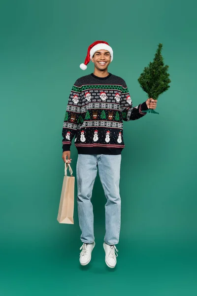 Piena lunghezza di felice afro americano uomo in cappello di Babbo Natale levitante con shopping bag e piccolo albero di Natale su sfondo verde — Foto stock