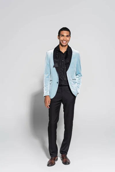Longitud completa del hombre afroamericano en chaqueta de seda y pantalones negros posando con la mano en el bolsillo sobre fondo gris - foto de stock