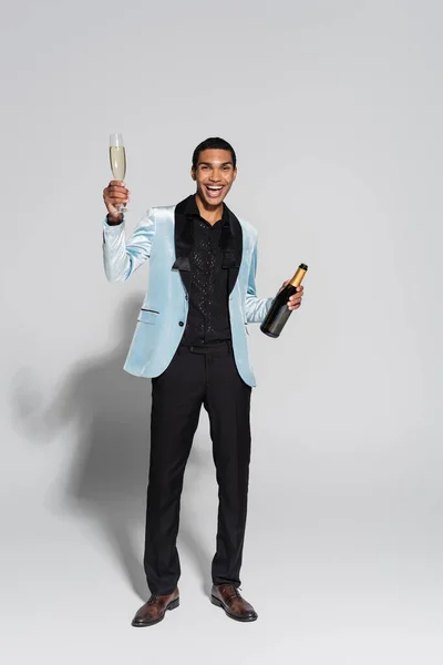 Longitud completa de hombre afroamericano elegante con botella y copa de champán sobre fondo gris - foto de stock