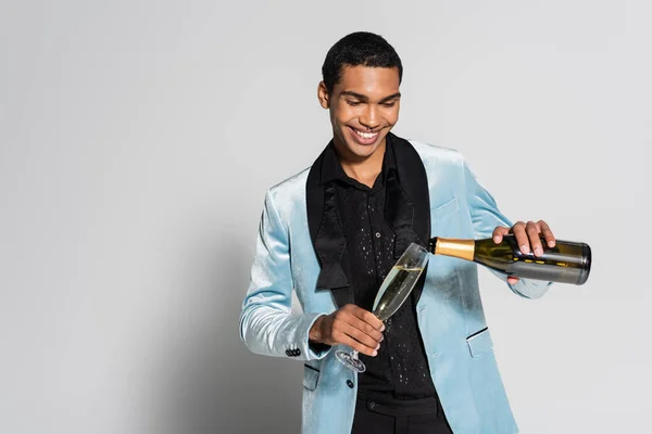 Fröhlicher afrikanisch-amerikanischer Mann in eleganter Seidenjacke, der Champagner auf grauem Hintergrund gießt — Stockfoto