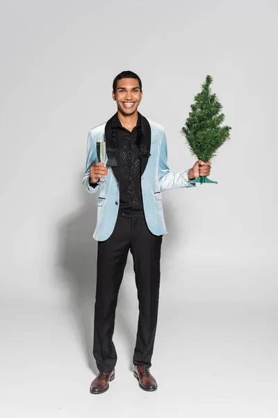 Alegre hombre afroamericano en ropa elegante sosteniendo pequeño árbol de Navidad y champán mientras sonríe a la cámara aislado en gris - foto de stock