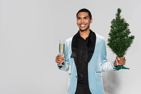 Элегантный африканский американец с бокалом шампанского и маленькой рождественской елкой, улыбающейся и смотрящей в сторону на сером фоне — стоковое фото