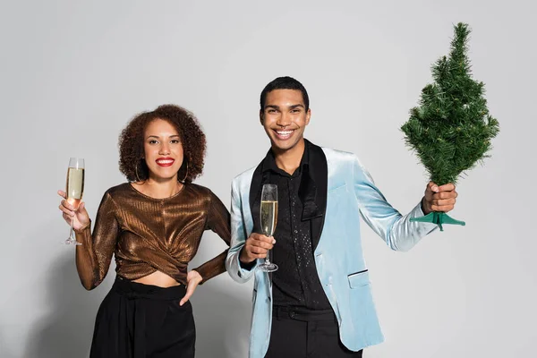 Elegante pareja afroamericana con copas de champán y pequeño árbol de Navidad sobre fondo gris - foto de stock
