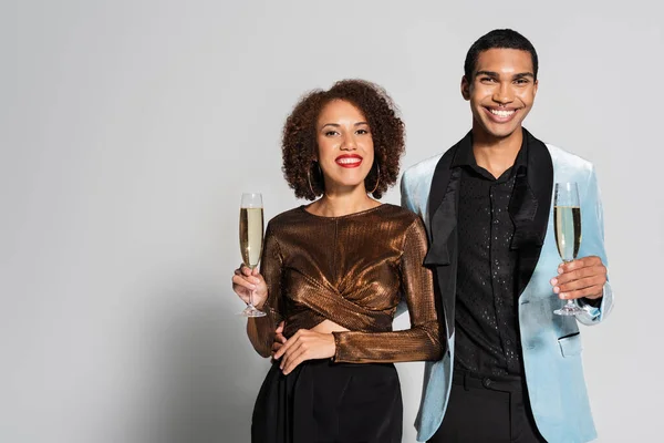 Mujer afroamericana en blusa brillante y hombre en chaqueta de seda sosteniendo copas de champán sobre fondo gris - foto de stock