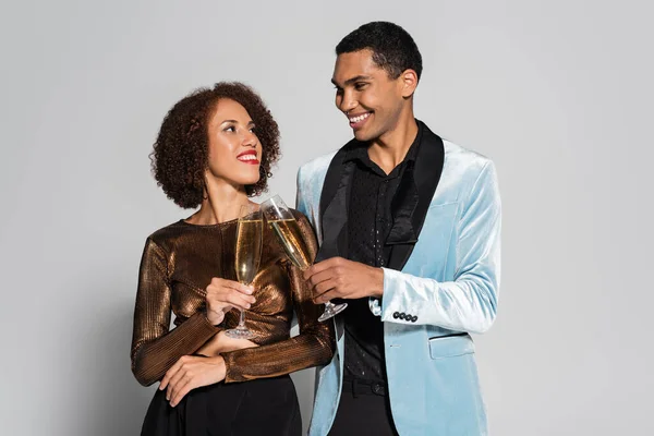 Молодая африканская американская пара в элегантной одежде звенящие бокалы шампанского и улыбающиеся друг другу изолированные на сером — стоковое фото