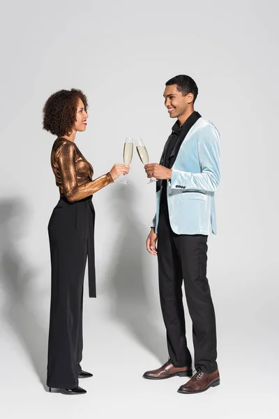 Вид сбоку на элегантную африканскую пару, держащую бокалы для шампанского и улыбающуюся друг другу на сером фоне — стоковое фото