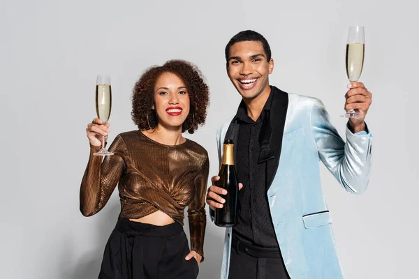 Feliz africano americano pareja en elegante ropa celebración de champán y mirando a la cámara en gris fondo - foto de stock