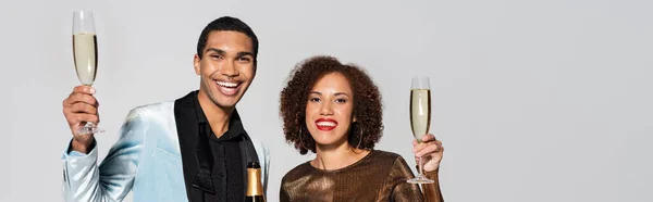 Стильная и элегантная африканская пара с бокалами для шампанского, улыбающаяся в камеру, изолированную на сером, баннере — стоковое фото