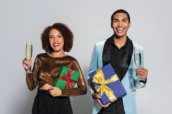 Felice coppia afro-americana in abiti alla moda con champagne e scatole regalo isolate sul grigio — Foto stock