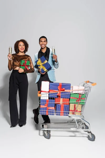 Элегантная африканская американская пара с бокалами шампанского и подарками, улыбающимися возле корзины с покупками на сером фоне — стоковое фото