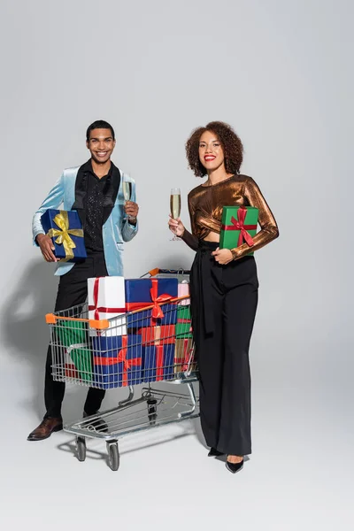Heureux couple afro-américain avec cadeaux et champagne debout près du panier avec des boîtes-cadeaux et sur fond gris — Photo de stock