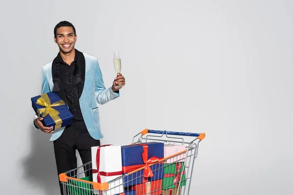 Stylish african american man with christmas present and champagne glass near shopping cart with gift boxes on grey background — Stock Photo