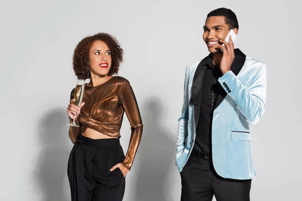 Homme afro-américain souriant en blazer de soie parlant sur smartphone près de femme élégante avec verre de champagne sur fond gris — Photo de stock