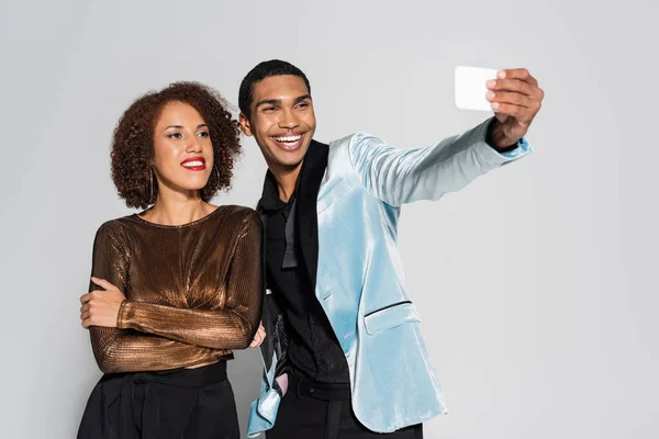 Alegre afroamericano hombre tomando selfie con elegante novia de pie con brazos cruzados aislado en gris - foto de stock