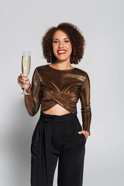 Femme afro-américaine en chemisier brun et brillant posant à la main dans la poche et verre de champagne isolé sur gris — Photo de stock