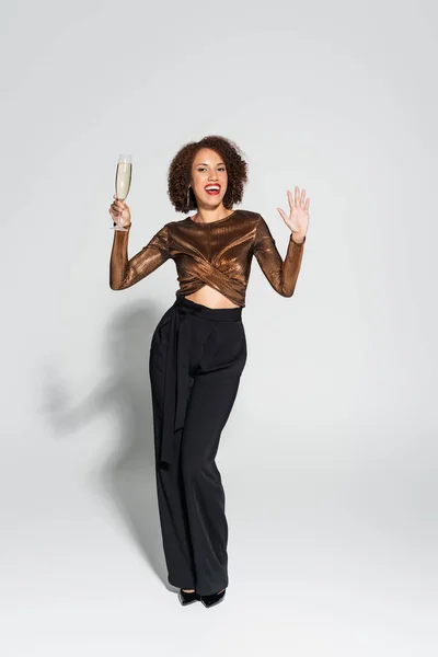 Full length of joyful and stylish african american woman holding champagne and waving hand on grey background — Stock Photo