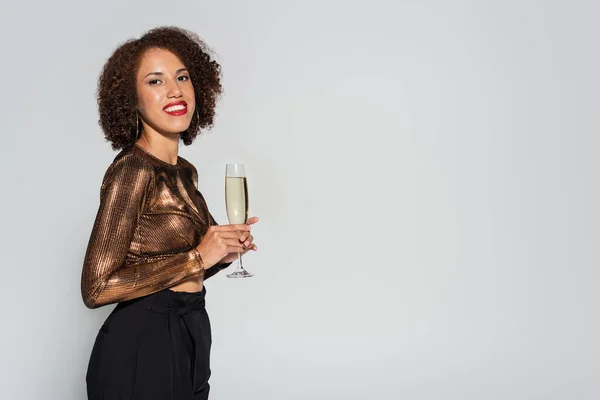 Curly african american woman in elegant shiny blouse smiling at camera while standing with champagne isolated on grey — Stock Photo