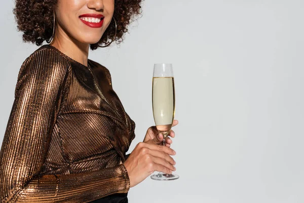 Vista recortada de mujer afroamericana elegante y sonriente con labios rojos sosteniendo copa de champán aislada en gris - foto de stock