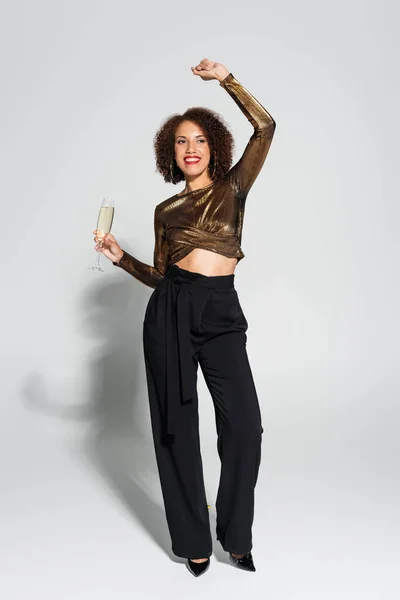 Pleine longueur de femme afro-américaine en chemisier marron et pantalon noir dansant avec verre de champagne sur fond gris — Photo de stock