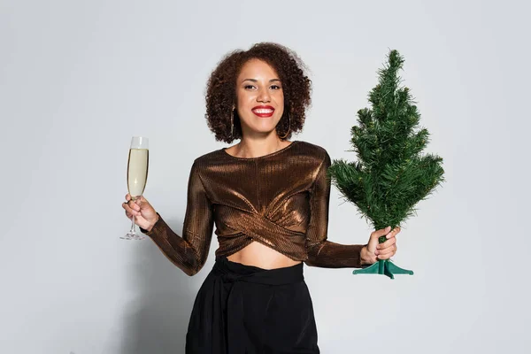 Elegante mujer afroamericana con pequeño árbol de Navidad y copa de champán sonriendo a la cámara sobre fondo gris - foto de stock