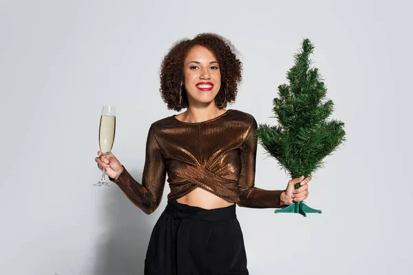 Stylish african american woman with small christmas tree and champagne glass smiling at camera on grey background — Stock Photo