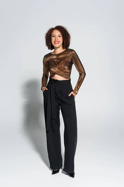 Curly african american woman in brown and shiny blouse posing with hands in pocket of black pants on grey background — Stock Photo