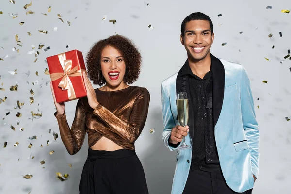 Erfreut afrikanisch-amerikanische Frau mit Weihnachtsgeschenk in der Nähe eleganter Mann mit Champagnerglas auf grau — Stockfoto