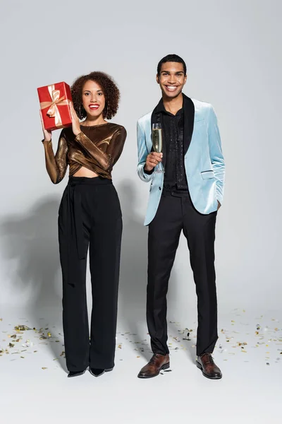 Elegante afrikanisch-amerikanische Mann mit Champagnerglas und Hand in Tasche in der Nähe Freundin mit Weihnachtsgeschenk auf grauem Hintergrund — Stockfoto