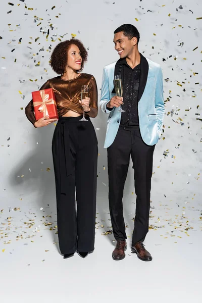 Élégant couple afro-américain avec champagne et coffret cadeau se souriant sur gris — Photo de stock