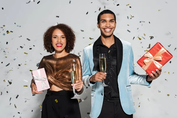 Alegre y elegante pareja afroamericana con regalos y champán sonriendo a la cámara sobre fondo gris - foto de stock