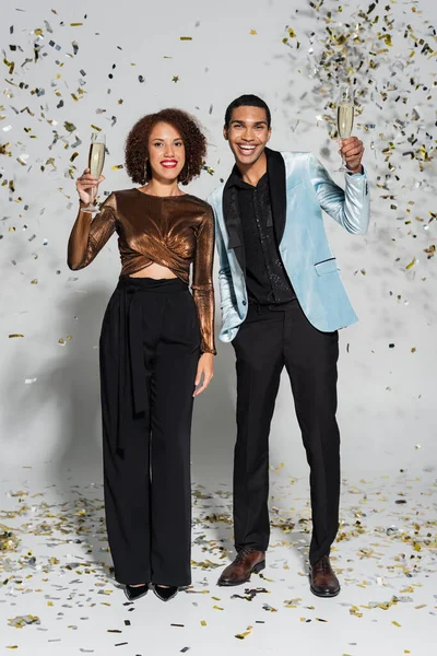 Heureux couple afro-américain en vêtements de fête tenant champagne près de confettis sur gris — Photo de stock