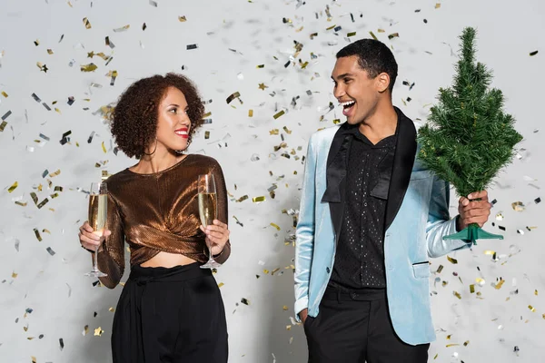 Sonriente hombre afroamericano con pequeño árbol de navidad mirando a mujer elegante con copas de champán sobre fondo gris - foto de stock