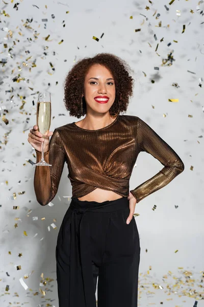 Joyeuse femme afro-américaine debout avec la main sur la hanche et verre de champagne près de confettis sur fond gris — Photo de stock