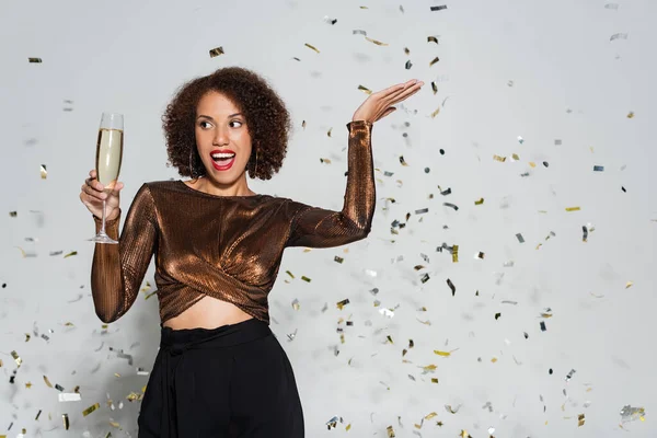 Mujer afroamericana emocionada en ropa festiva posando con copa de champán cerca de confeti sobre fondo gris - foto de stock