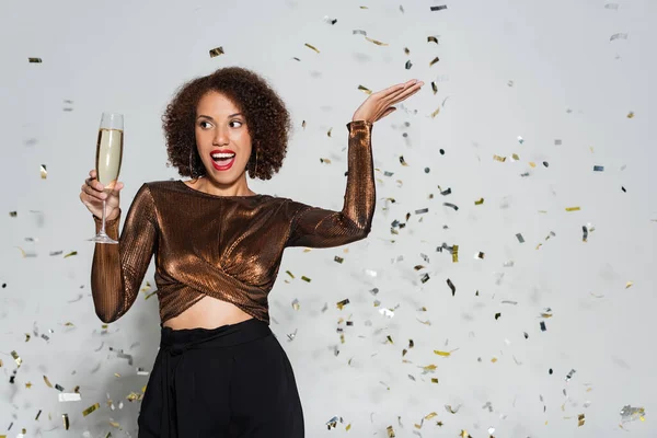 Excitada afro-americana em roupas festivas posando com taça de champanhe perto de confete em fundo cinza — Fotografia de Stock