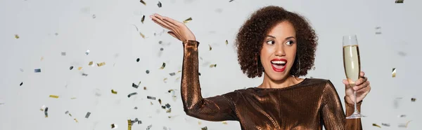 Aufgeregte und elegante afrikanisch-amerikanische Frau mit Champagnerglas, die neben Konfetti auf grauem Hintergrund posiert, Banner — Stockfoto