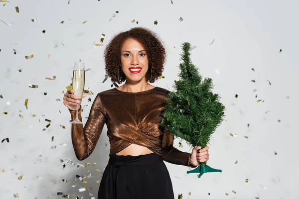 Sonriente mujer afroamericana en blusa brillante de pie con pequeño árbol de navidad y copa de champán cerca de confeti sobre fondo gris - foto de stock