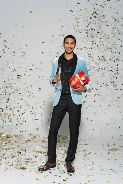 Cheerful african american man in elegant clothes standing with christmas present and champagne glass near confetti on grey — Stock Photo