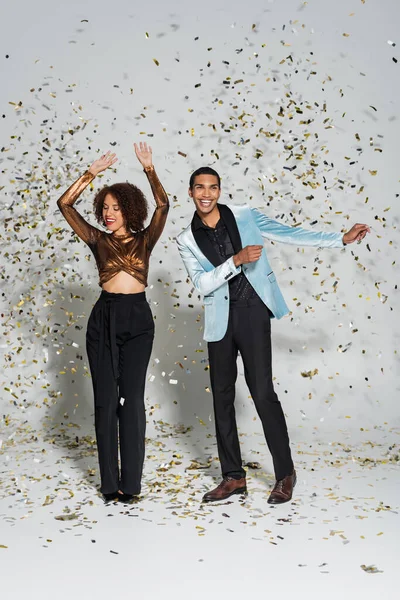 Pareja afroamericana emocionada y elegante bailando durante la fiesta de Navidad cerca de confeti espumoso en gris - foto de stock